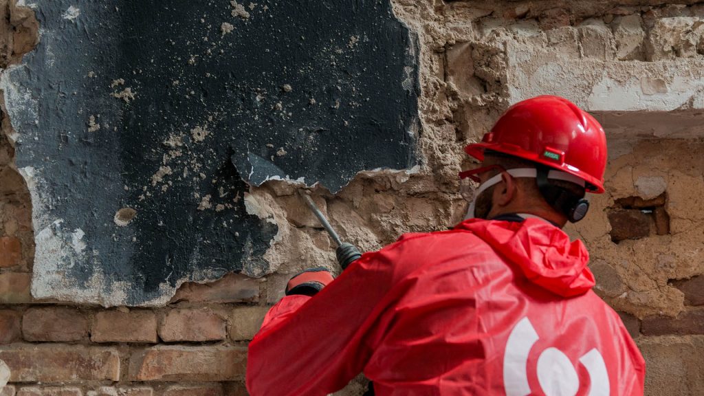 Ein BELFOR-Mitarbeiter in einem roten PSA-Anzug entfernt eine beschädigte Wand