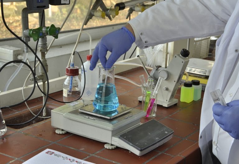 Une main gantée tient un agitateur en verre dans un erlenmeyer rempli de liquide bleu sur une balance électronique