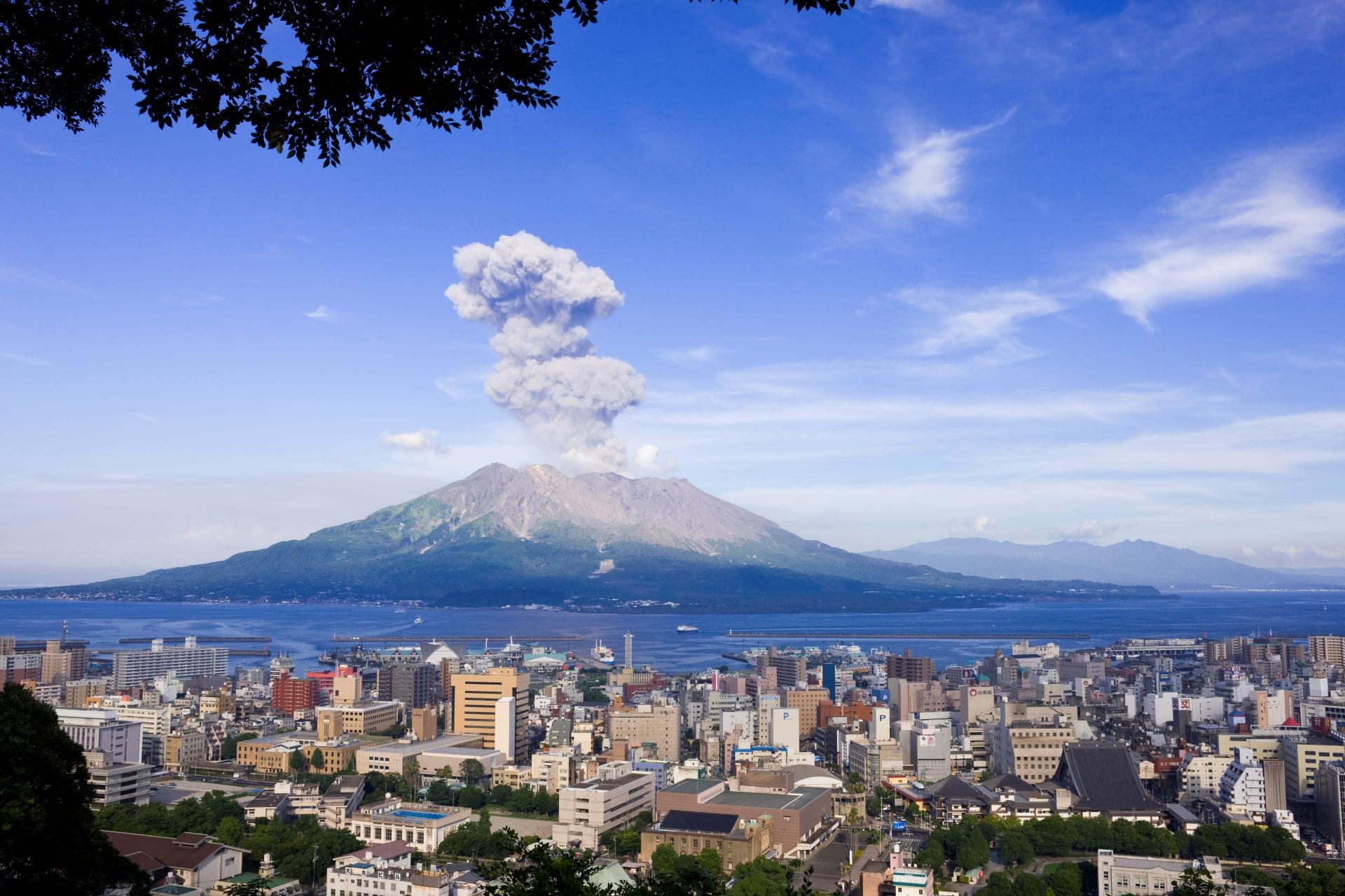 View of a city with a volcano in the background erupting, sending a plume of smoke into the clear blue sky