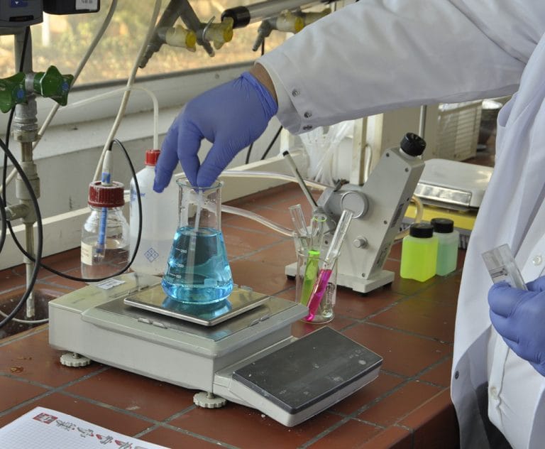 Gloved hand holding a glass stirrer in a conical flask filled with blue liquid on an electronic balance