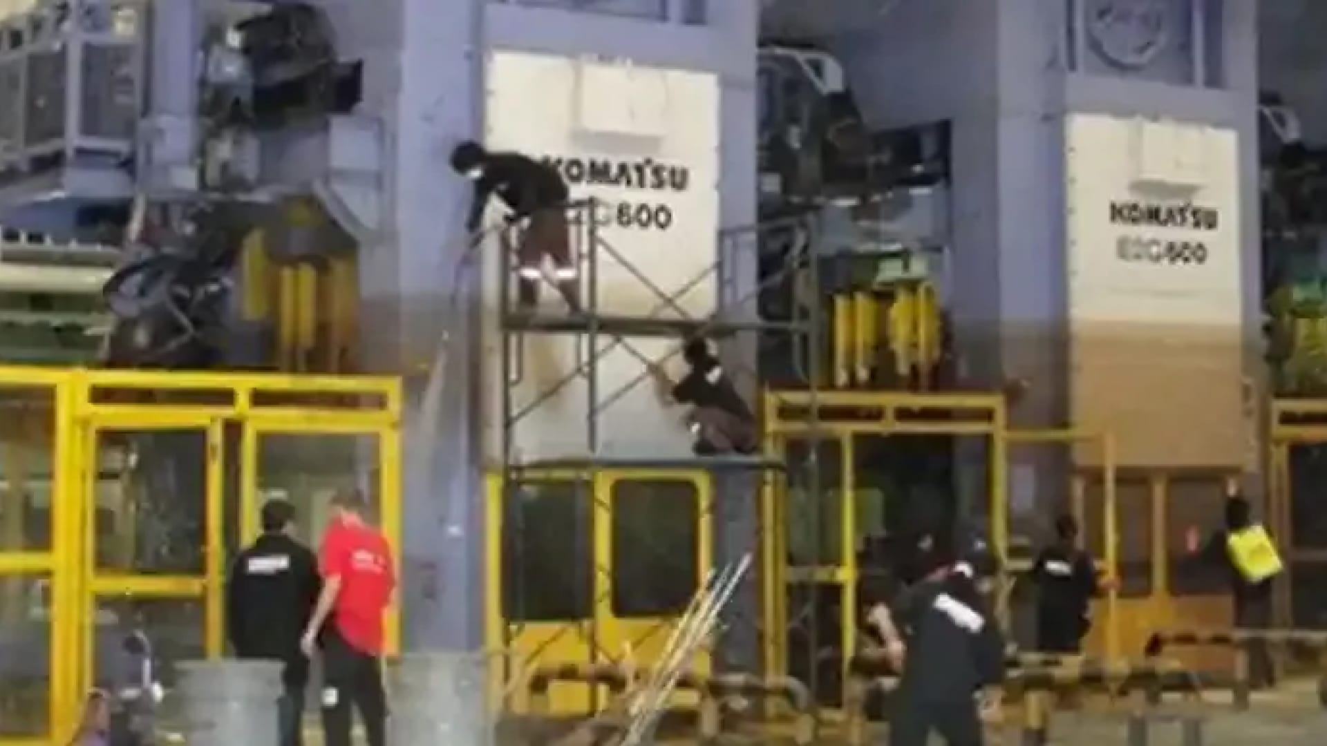 Workers assembling or maintaining large industrial machinery, with two individuals on scaffolding and others on the ground near Komatsu equipment