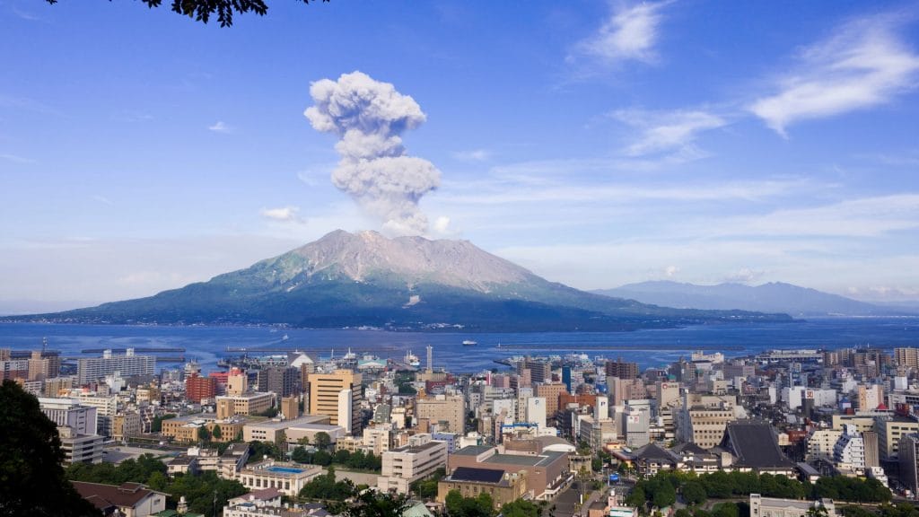 View of a city with a volcano in the background erupting, sending a plume of smoke into the clear blue sky